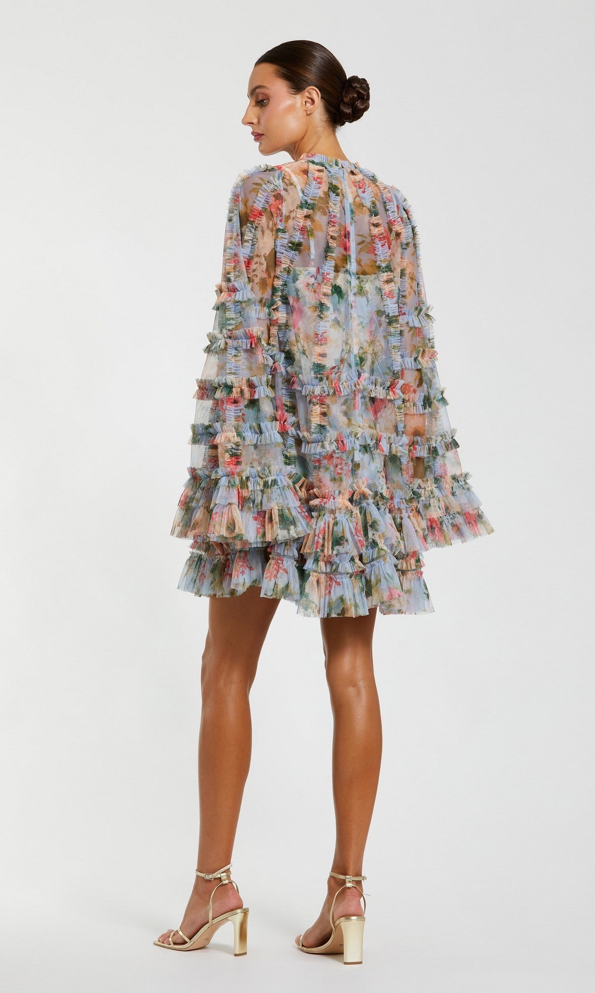 A woman stands in profile wearing the Mac Duggal 8300 Bell-Sleeve Floral Wedding Guest Dress, featuring sheer floral fabric, tiered ruffles, and bell sleeves, paired with strappy high-heeled sandals.